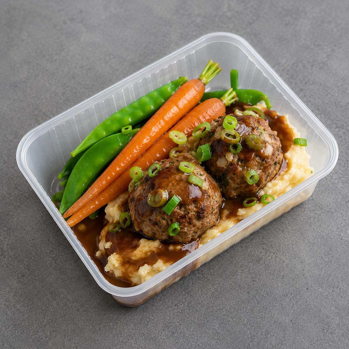 A clear plastic container holding a prepared meal with two browned meatballs on mashed potatoes, topped with green onions and brown gravy, served with orange carrots and green snap peas on a gray surface.