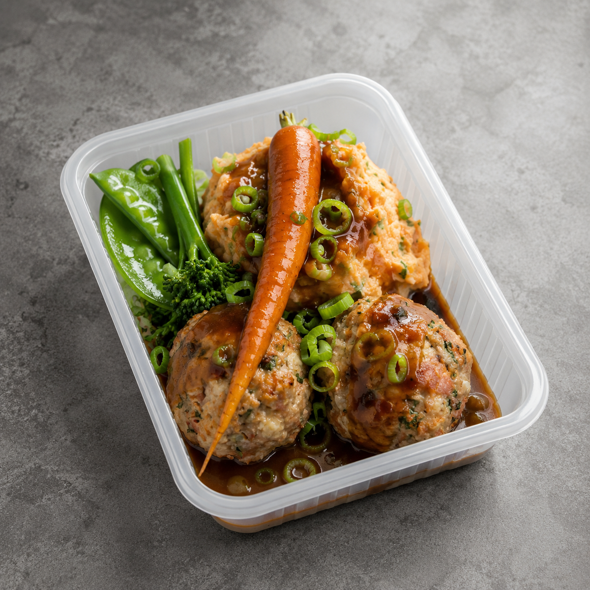 A clear plastic container holding a prepared meal with two meatballs in brown sauce, mashed potatoes topped with sliced green onions, a whole carrot, snap peas, and broccoli on a gray surface.