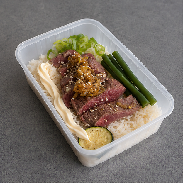 A clear plastic container filled with a prepared rice bento box on a gray surface. The meal includes sliced beef, white rice, green onions, green beans, a squeeze of mayonnaise, a cucumber slice, and a brown sauce topping with sesame seeds.
