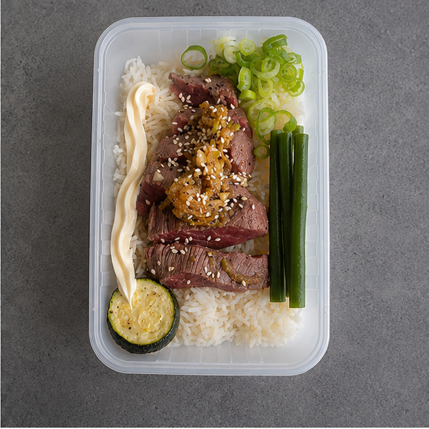 A clear plastic bento container filled with sliced beef on white rice, topped with sesame seeds and a sauce drizzle, served with sliced green onions and a zucchini on a gray surface.